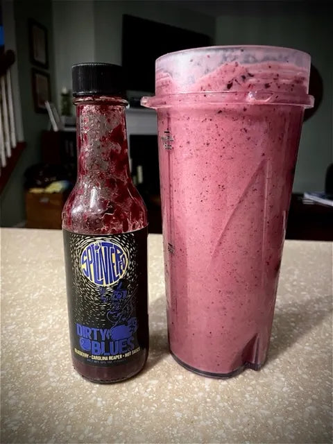 Bottle of Splintered Dirty Blues hot sauce Blueberry juice next to a pink smoothie in a clear container on a countertop.