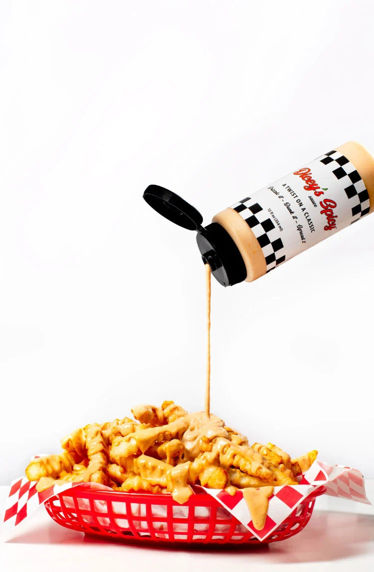 A bottle of Dippy's Burgers creamy cheese sauce is tilted to pour a stream of golden yellow sauce over a serving of waffle fries in a red basket lined with red and white checkered paper, on a white background.