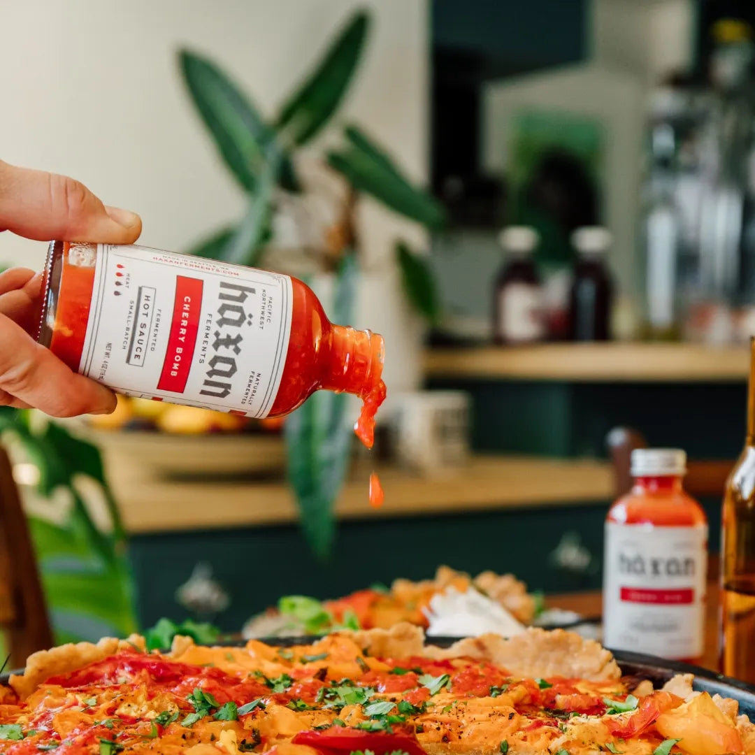 Person pouring sauce from a bottle labeled 'haxan' onto a pizza in a kitchen setting.