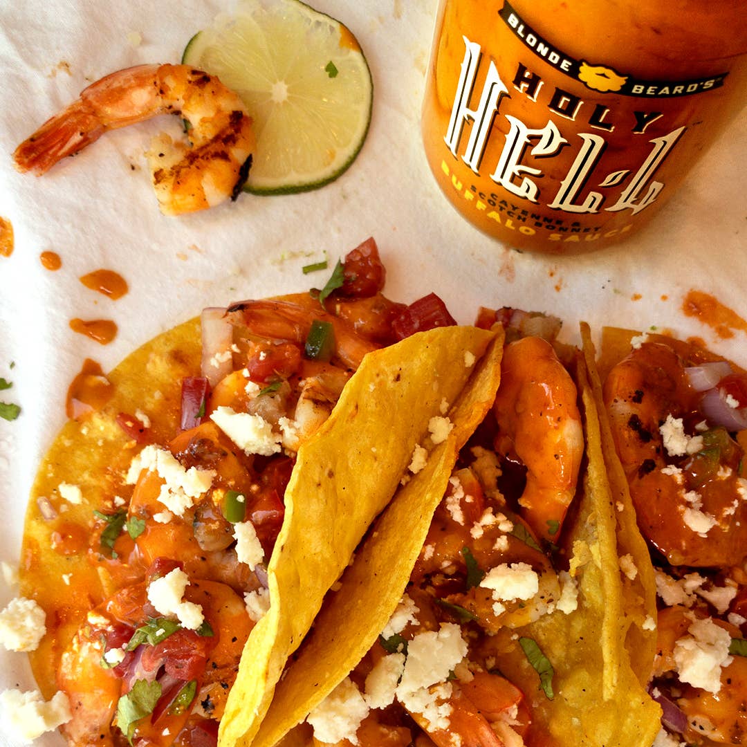 Tacos with shrimp and lime slices on a tortilla, accompanied by a bottle of 'Holy Hell' hot sauce.