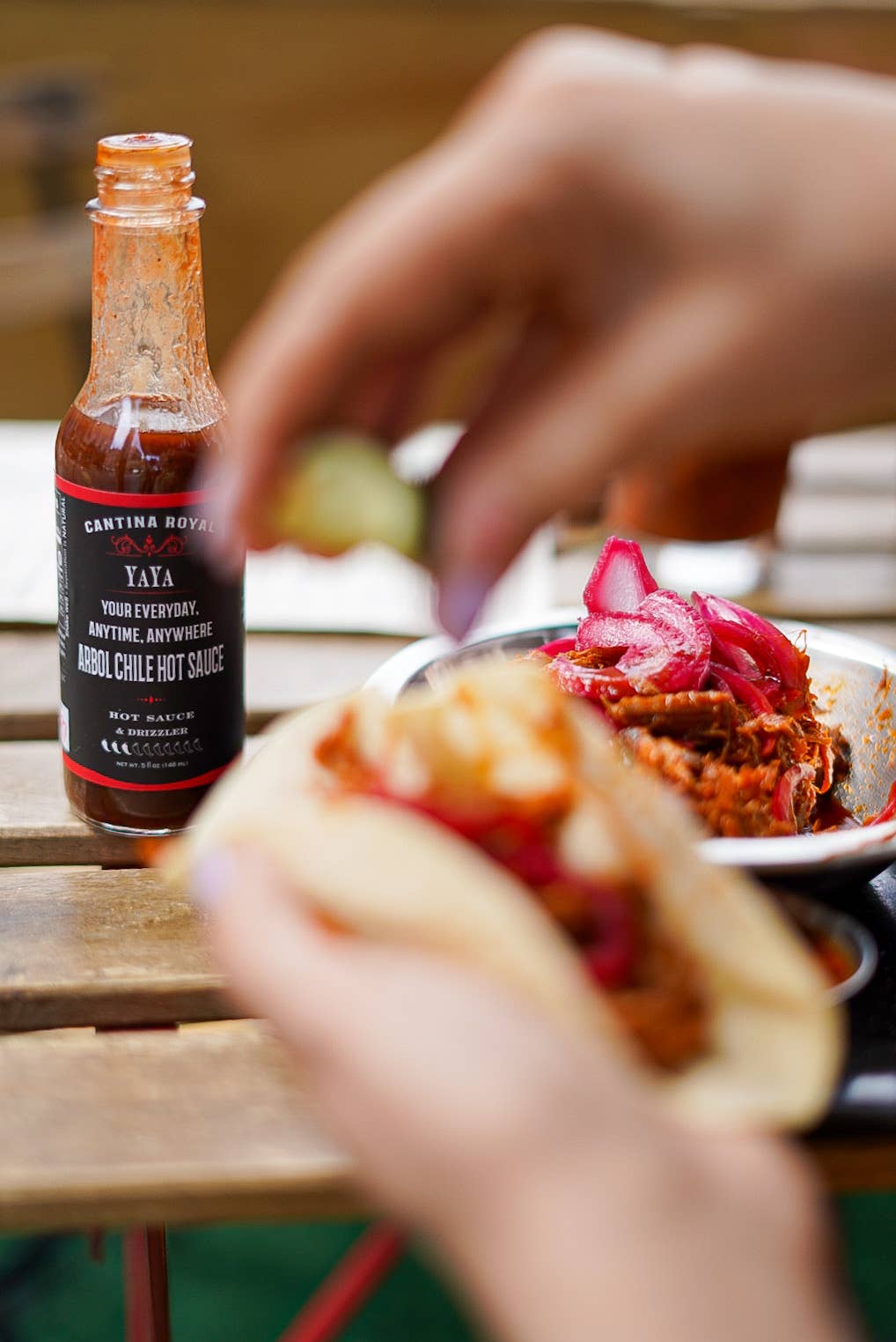 Person holding a sandwich with a bottle of hot sauce on a table