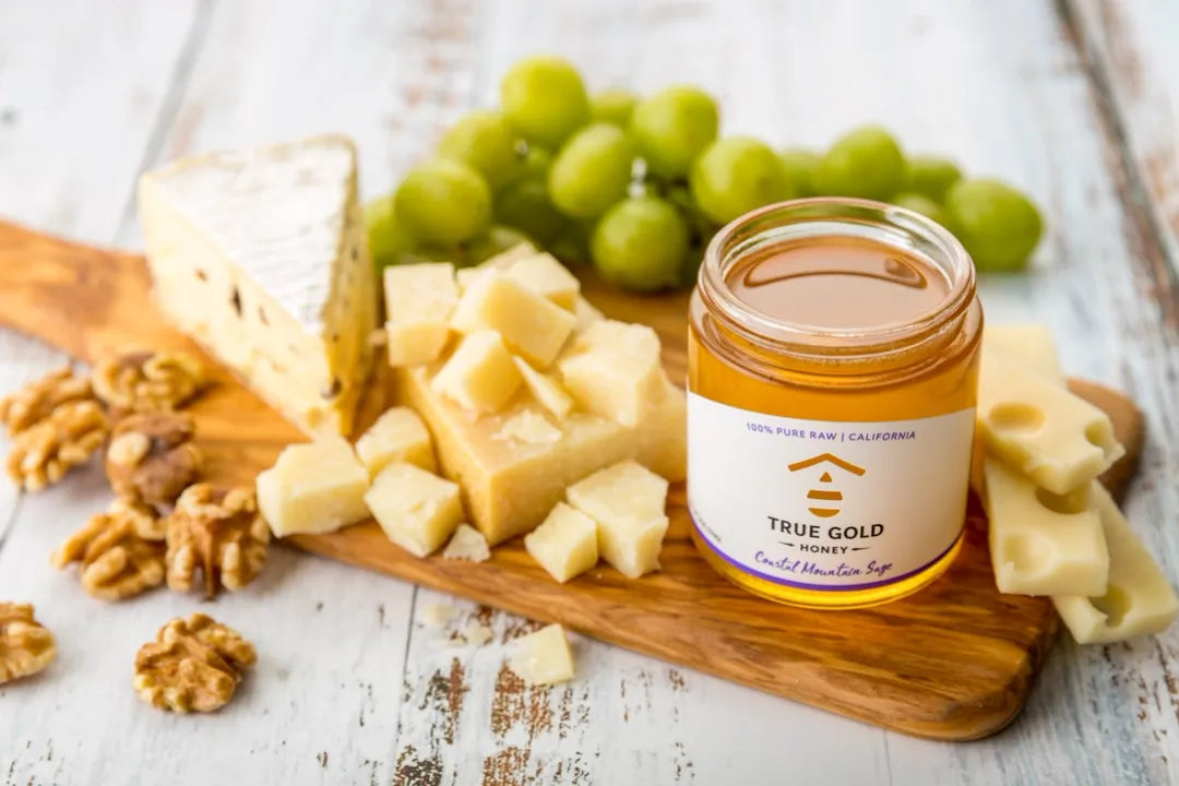 Jar of 'True Gold' honey on a wooden board with cheese, grapes, and nuts.