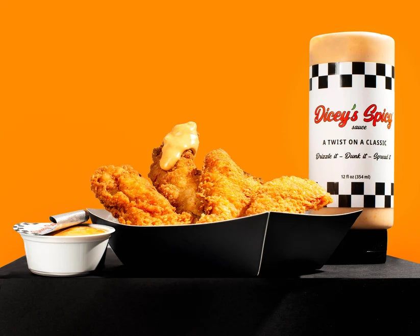 A food product display featuring golden-brown fried chicken pieces in a black tray, a small white dipping sauce cup with a silver spoon, and a tall bottle of Dice's Spicy Sauce against a solid orange background.