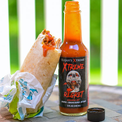 Bottle of Elijah's Extreme Regret hot sauce next to a wrapped burrito on a table with a green outdoor background.