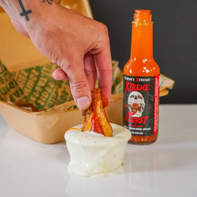 Person dipping a chicken wing into a bowl of sauce with a bottle of hot sauce in the background.
