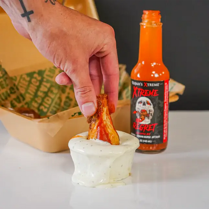 Person dipping a chicken wing into a bowl of sauce with a bottle of hot sauce in the background.