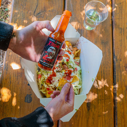 Person holding a bottle of hot sauce over a colorful salad on a wooden table.