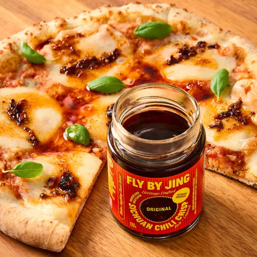 Jar of 'Fly By Jing' chili crisp sauce next to a pizza on a wooden surface