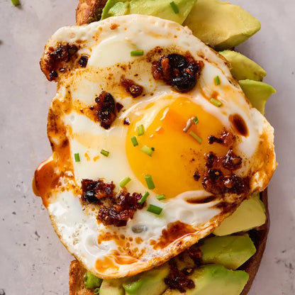 Fried egg on toast with extra spicy chili crisp and avocado slices on a gray surface