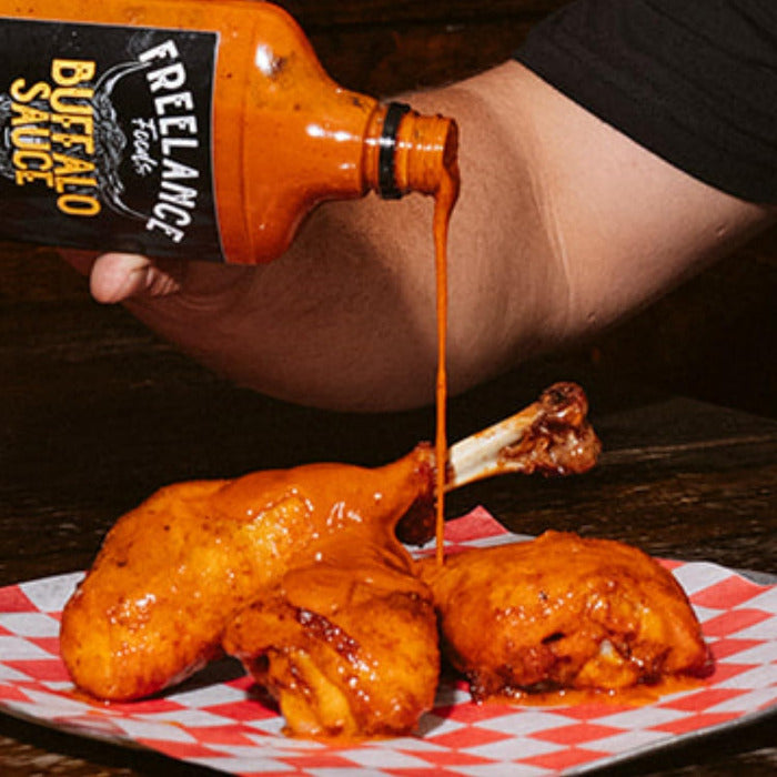 Person pouring Freelance Buffalo Sauce onto fried chicken on a checkered paper.