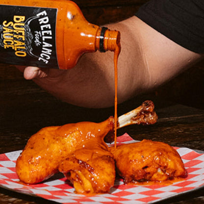 Person pouring Freelance Buffalo Sauce onto fried chicken on a checkered paper.