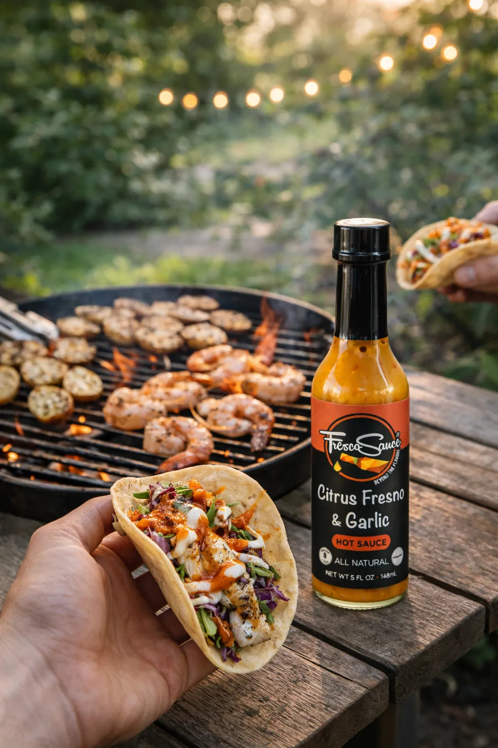 Person holding a taco with a bottle of Tito's Curly Fresno & Garlic hot sauce on a grill and outdoor setting.