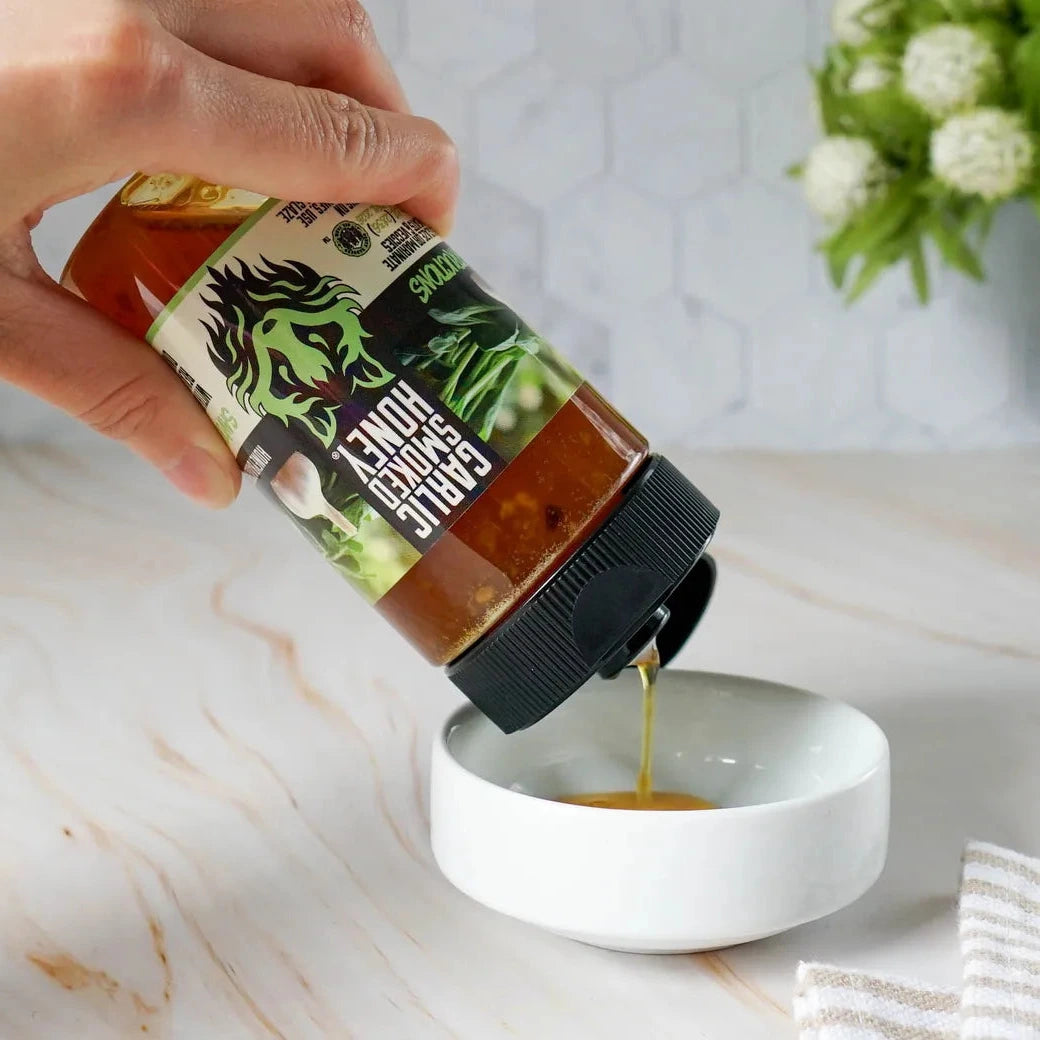 Hand pouring honey from a jar labeled 'Honey Sticks' into a white bowl on a marble surface.