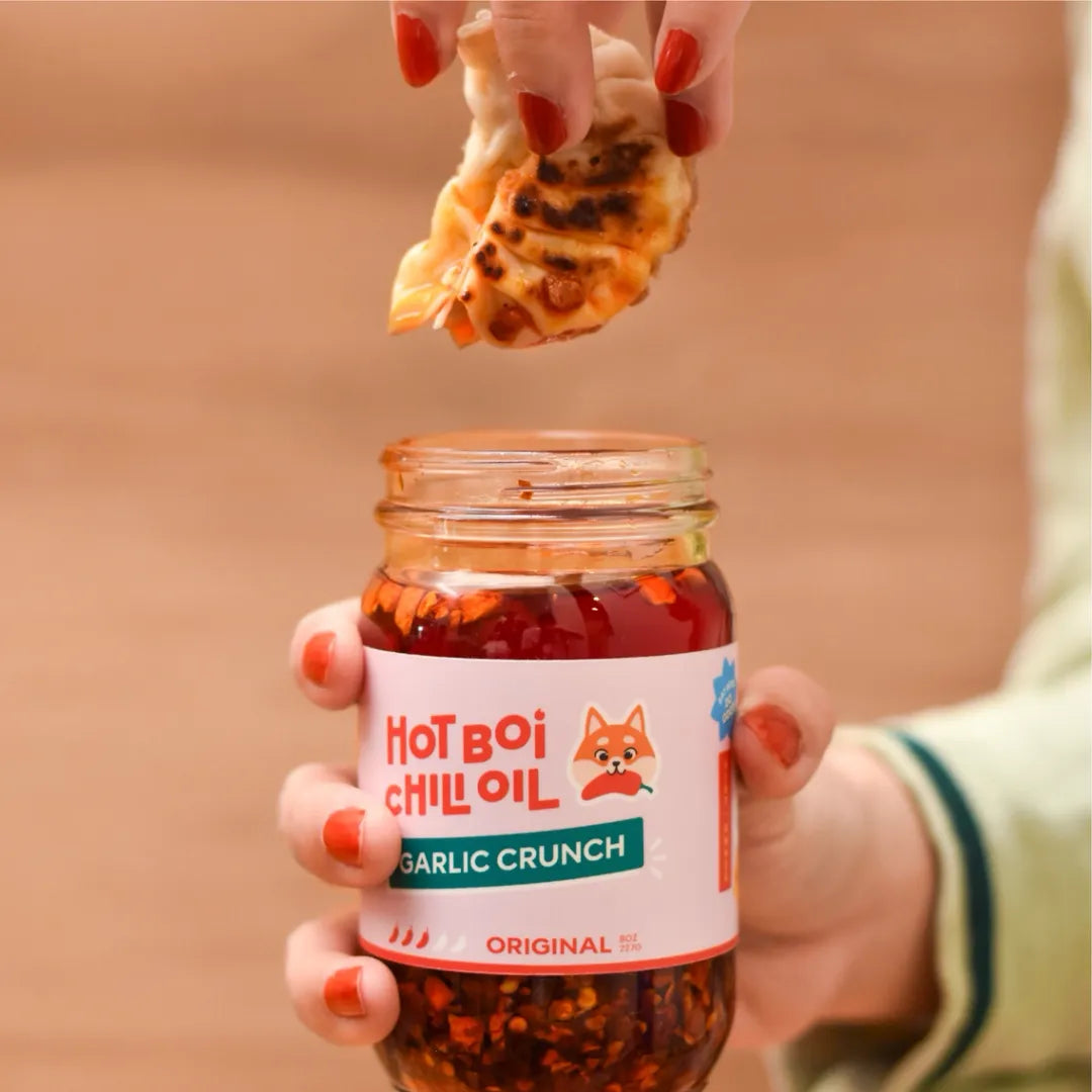 Hand holding a jar of 'Hot Boi Chilli Oil Garlic Crunch' with a piece of bread being dipped into it.