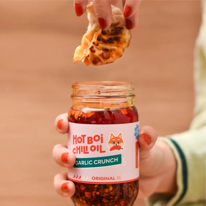 Hand holding a jar of 'Hot Boi Chilli Oil Garlic Crunch' with a piece of bread being dipped into it.