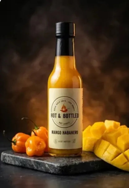 Bottle of Hot & Bottled Mango Habanero hot sauce with habanero peppers and mango on a dark background