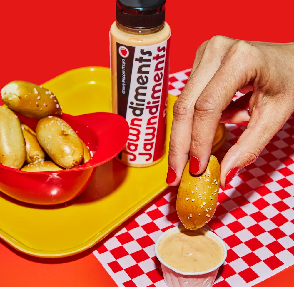 Person dipping a pretzel into a plastic cup of Jawndiments cherry mayo on a red and white checkered tablecloth