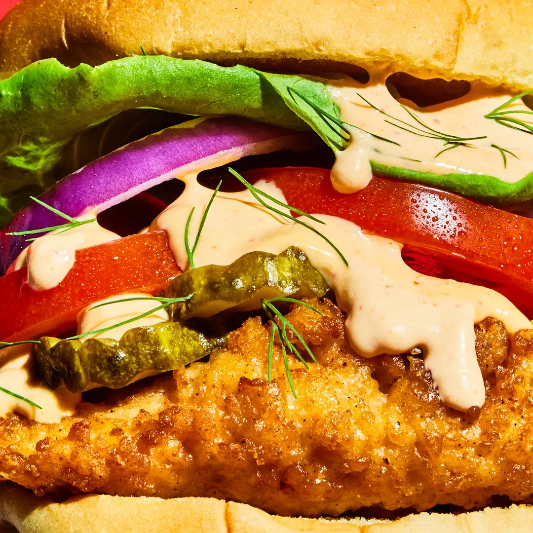 Close-up of a sandwich with fried fish, lettuce, tomato, and pickles on a bun.
