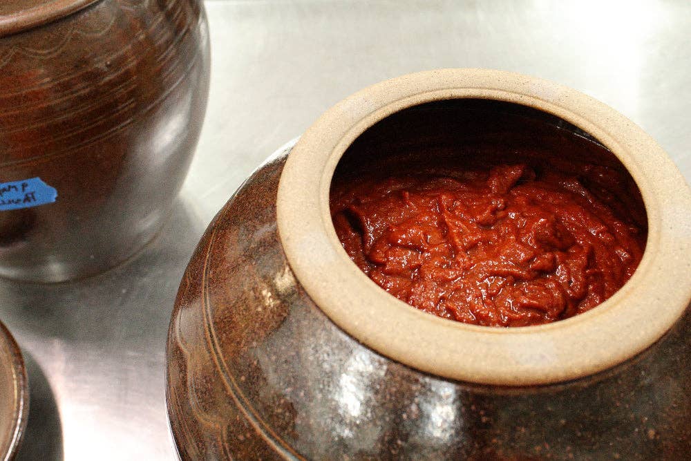 A close-up of a dark brown ceramic Korean jar filled with red kimchi, set on a metallic surface next to other jars.