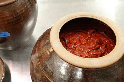 A close-up of a dark brown ceramic Korean jar filled with red kimchi, set on a metallic surface next to other jars.