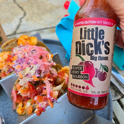 Bottle of Little Dick's Reaper Cherry Bourbon hot sauce with tacos in the background