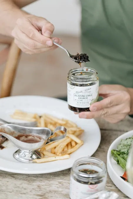 Person holding a jar of jalapeno honey chili crisp with a spoon over a plate of food.