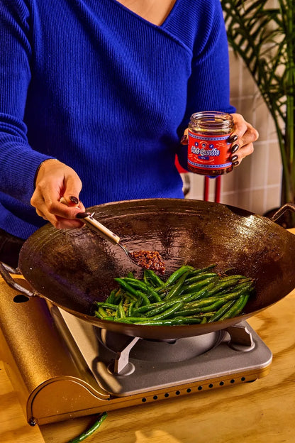 Person cooking green beans in a wok with a jar of sauce on a stove.