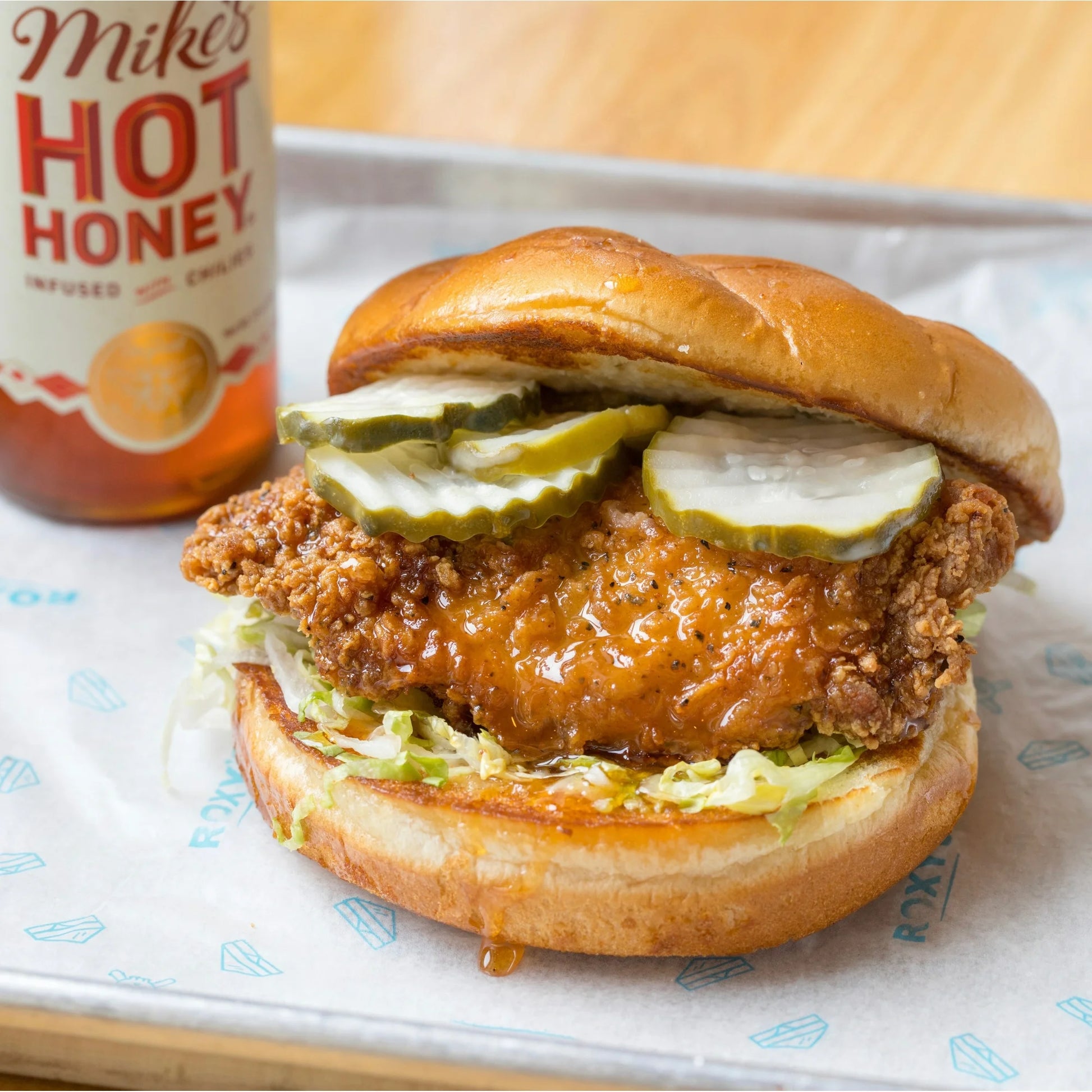 Burger with fried chicken, pickles, and lettuce on a bun next to a bottle of Mike's Hot Honey.
