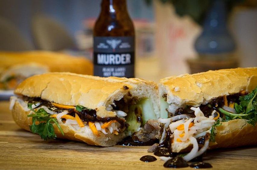 Sub sandwich cut in half on a wooden board with a bottle or Murder Hornet Black Label Hot Sauce in the background.