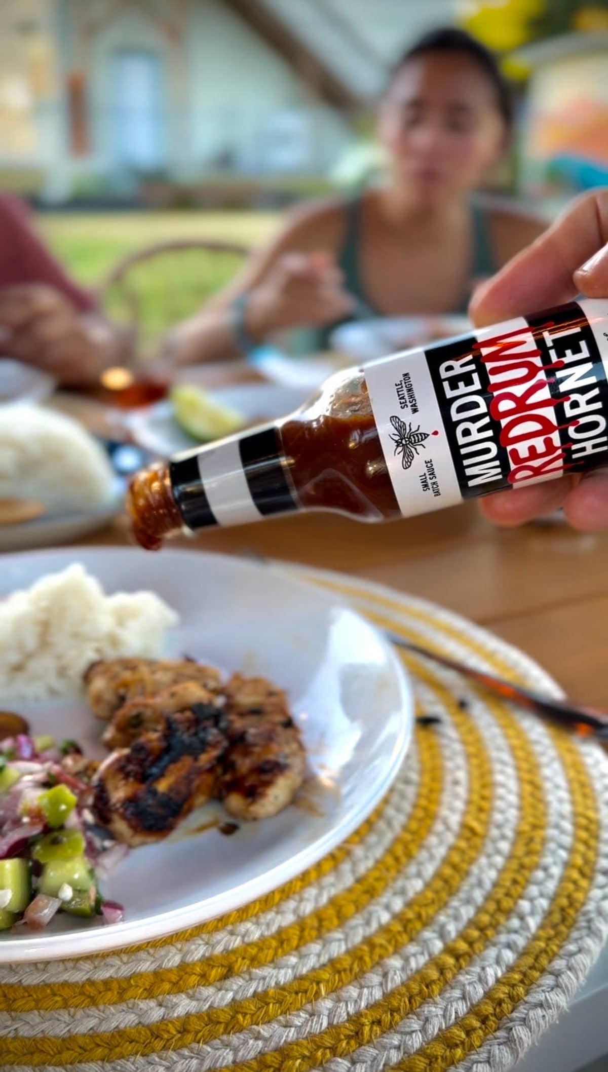 Person holding a bottle of 'Murdered Rum' sauce over a plate of food with a blurred background