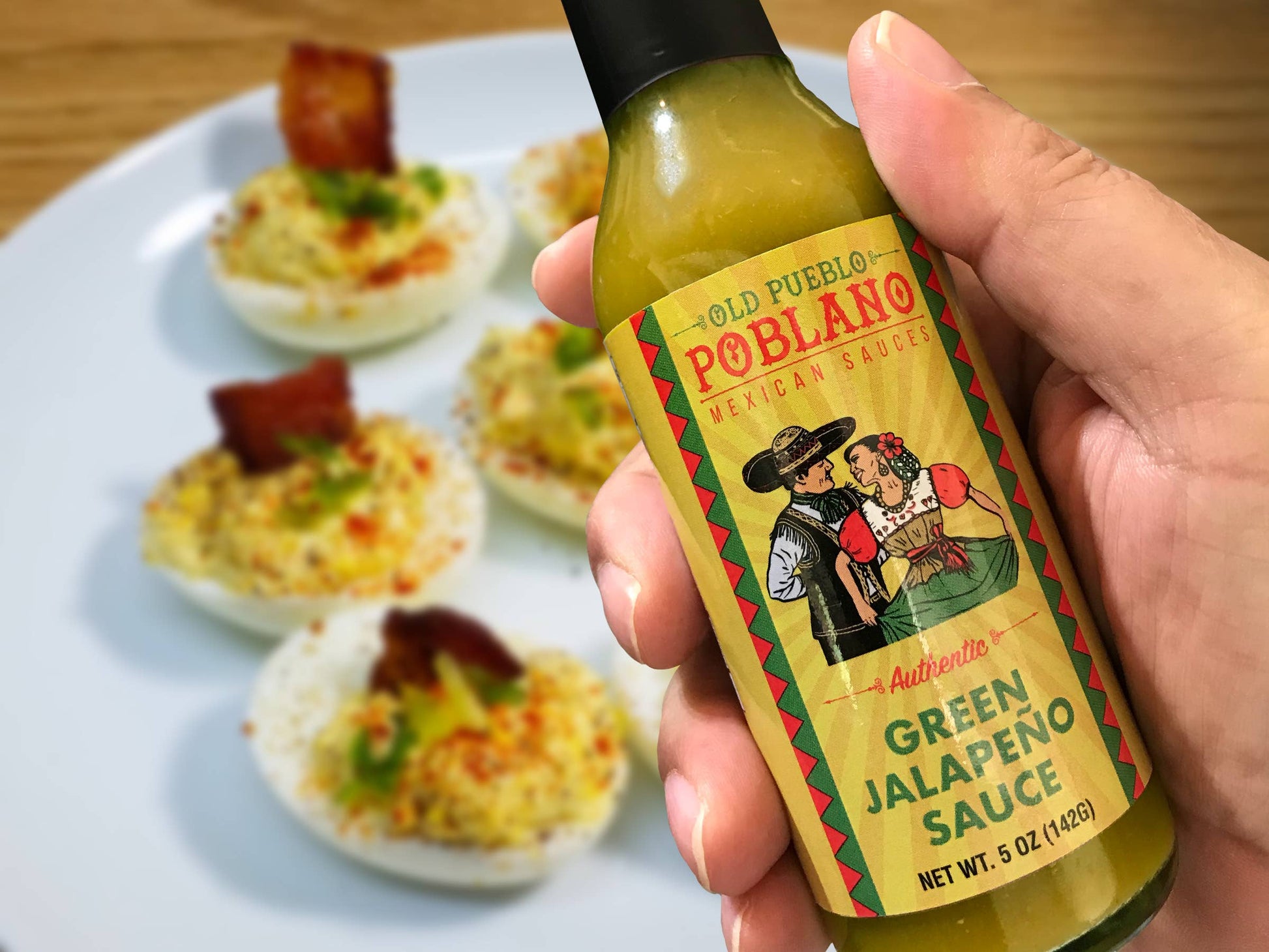 Hand holding a bottle of Old Pueblo Poblano Green Jalapeno Sauce with deviled eggs in the background.