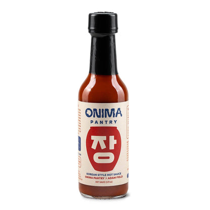A bottle of Onima Pantry Korean Style Hot Sauce on a solid white background. The bottle is made of clear glass showing the reddish-brown sauce inside, topped with a black cap and featuring a cream-colored label with blue and red text.