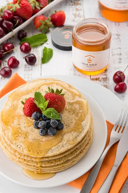 Stack of pancakes with berries on a plate, accompanied by a jar of honey and fruits.