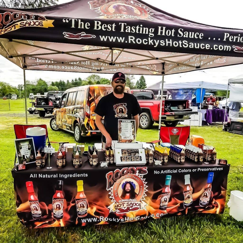 Person standing behind a Rocky's Hot Sauce display at an outdoor event.