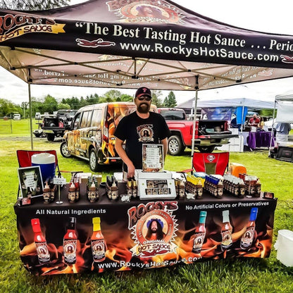 Person standing behind a Rocky's Hot Sauce display at an outdoor event.