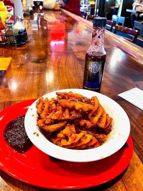 Breaded and fried food on a white plate with a red rim, served on a wooden table with Splintered Dirty Blues hot sauce.
