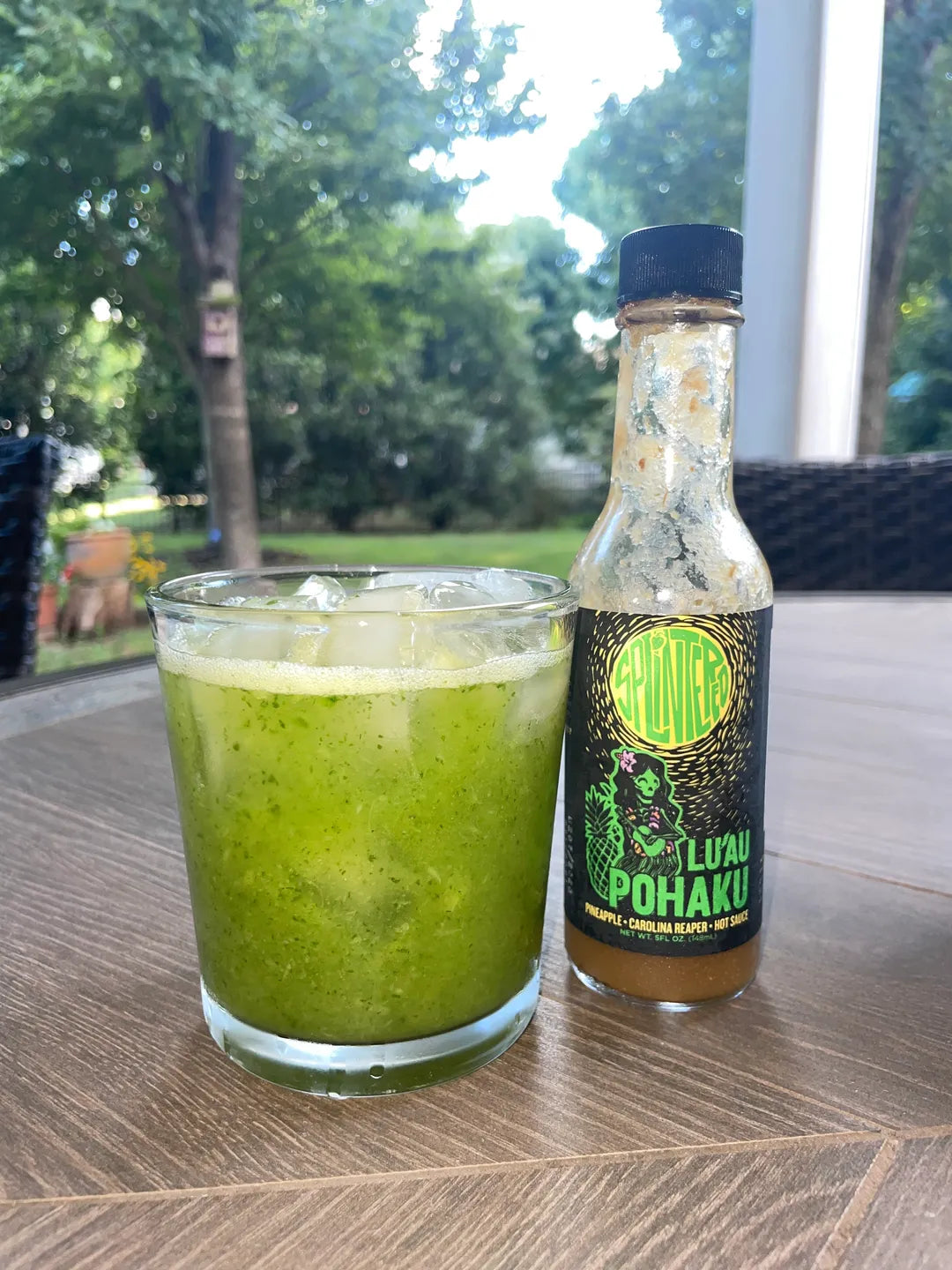 Green drink in a glass with a bottle of 'Fluai Pohaku' on a wooden table outdoors.