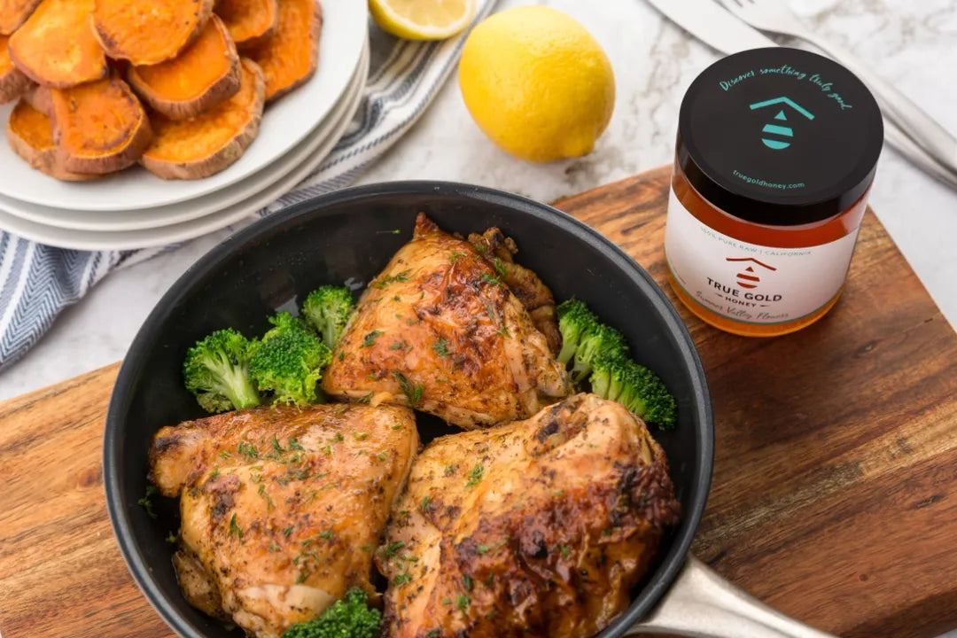 Baked chicken thighs with broccoli in a black dish on a wooden cutting board, accompanied by a jar of True Gold honey.