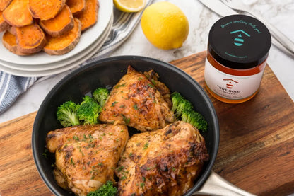Baked chicken thighs with broccoli in a black dish on a wooden cutting board, accompanied by a jar of True Gold honey.