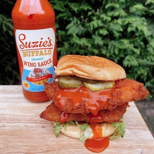 Burger with fried chicken, pickles, and sauce next to a bottle of Suzie's Buffalo Wing Sauce on a wooden surface.