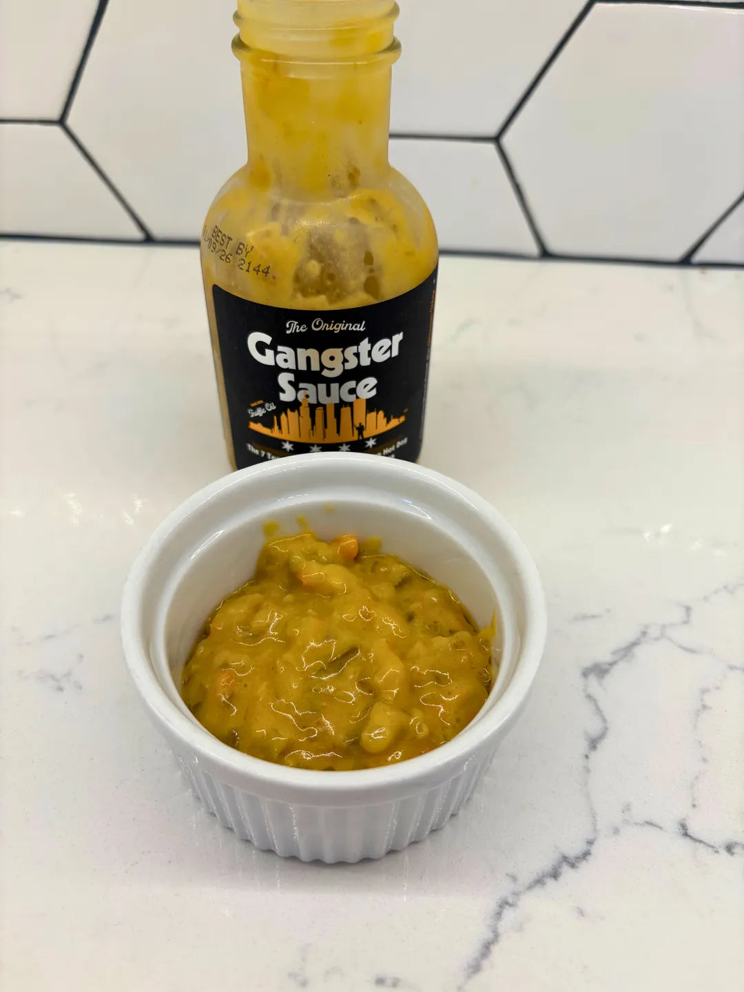 Bottle of 'Gangster Sauce' with a small container of sauce on a marble surface.
