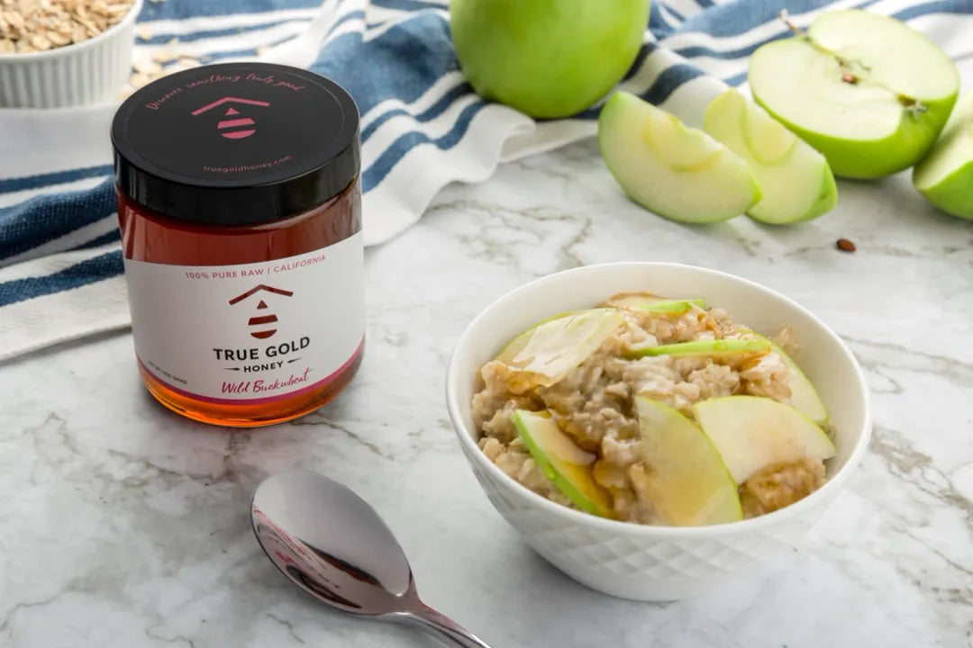 Jar of True Gold honey, bowl of oatmeal with apples, and sliced apples on a marble surface.