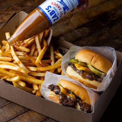 Two burgers and fries with a bottle of Zab's hot sauce on a wooden surface
