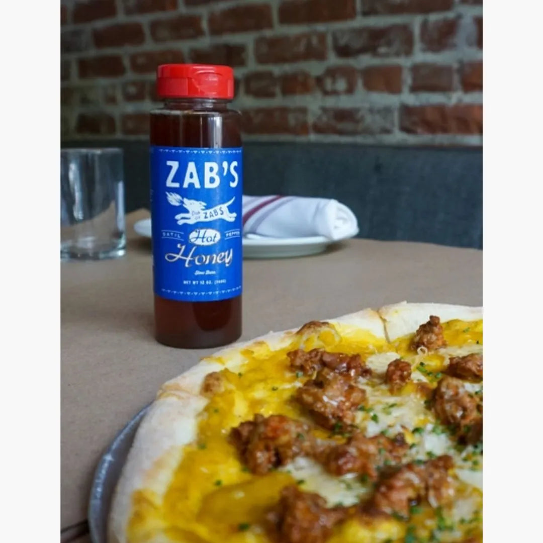 Bottle of Zab's Honey on a table with a pizza in the foreground, against a brick wall.