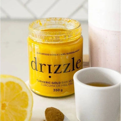 Jar of Drizzle Turmeric Gold honey with a lemon, cookie, and cup on a table.