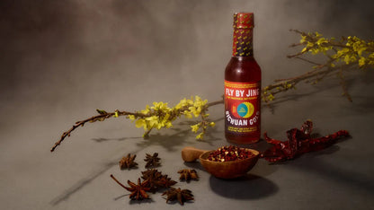 Bottle of Fly By Jim's hot sauce with spices and flowers on a gray background