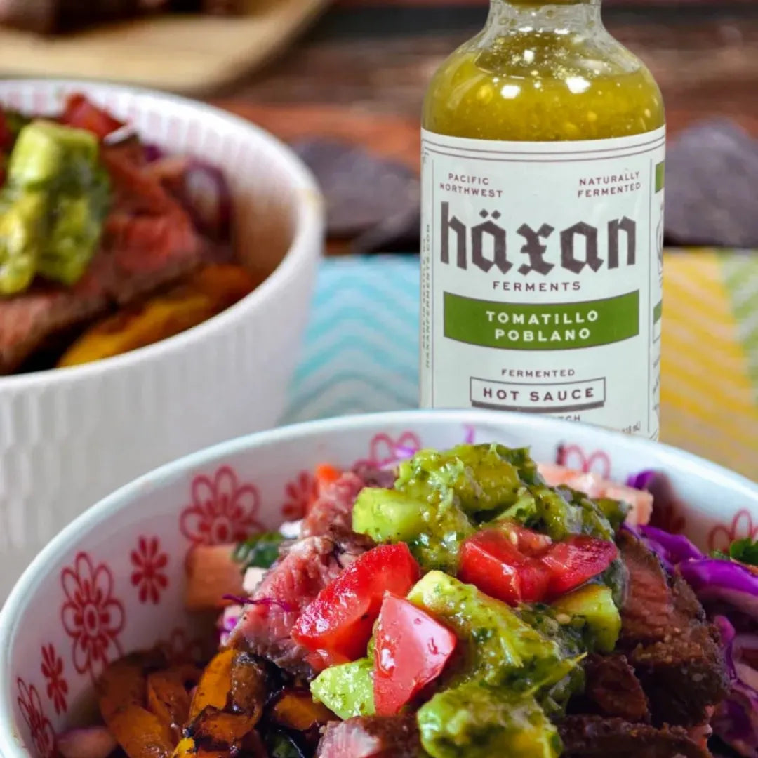 Bottle of Häxan Tomatillo Poblano hot sauce with a bowl of guacamole and meat dish in the background.