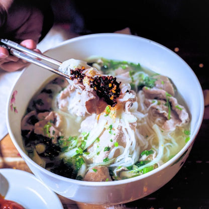 Bowl of noodles with vegetables and meat, topped with black pepper, on a wooden table.