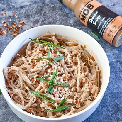 Bowl of noodles with green onions and a bottle of MOB Sesame Satay sauce on a gray surface.