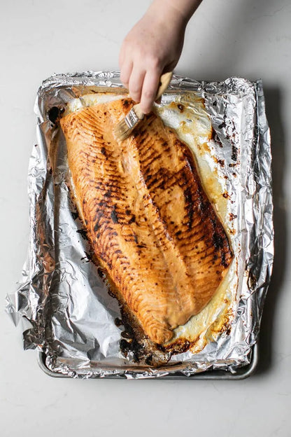 Hand holding a piece of grilled salmon on a foil-lined baking tray.